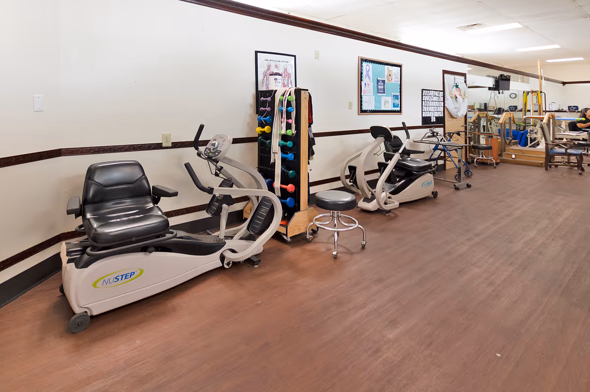 Interior exercise/physical therapy room with recumbent bikes, free weights, walkers and seating in a senior living facility.