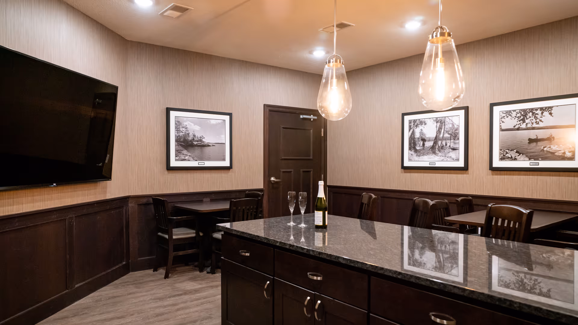 Interior room with a large granite countertop island in the foreground, two hanging pendant lights above it, a bottle of champagne and two champagne flutes on the countertop. The room has dark wood paneling on the lower walls, beige wallpaper above, three black and white framed photographs on the walls, a dark wooden door, several dark wooden chairs and tables, and a large flat-screen TV mounted on the wall.