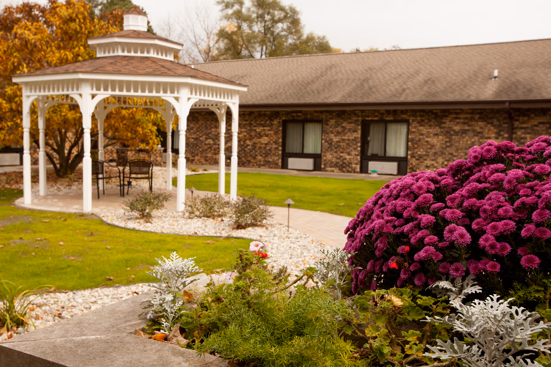 A well-maintained outdoor garden area at a senior living facility named White Oaks, featuring a white gazebo with chairs and a table underneath, surrounded by green grass, a stone pathway, and vibrant purple flowers in the foreground. The background shows a brick building with windows.