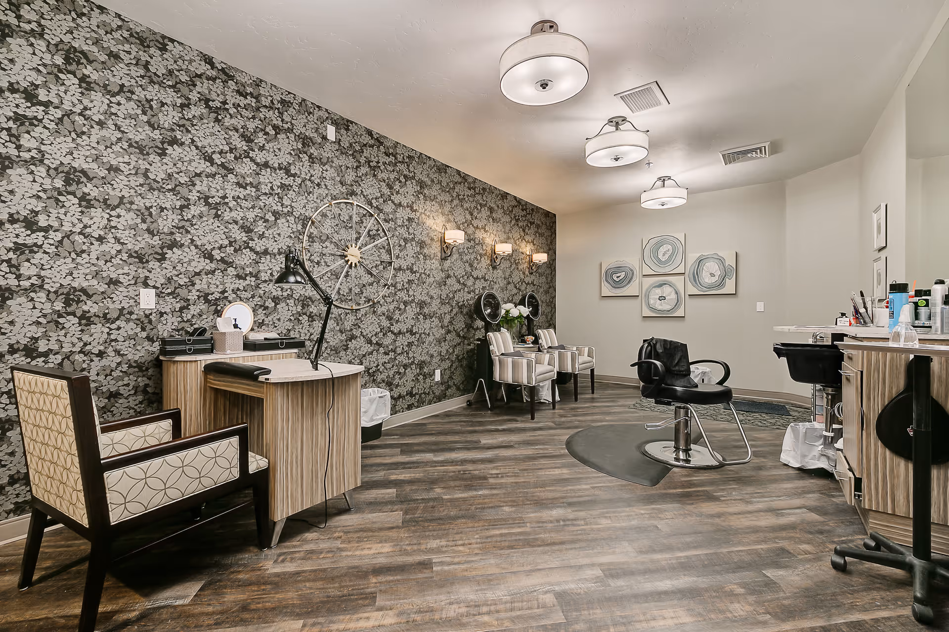 Interior view of a salon area in a senior living facility featuring a patterned accent wall, a styling chair, two hair drying chairs, a desk with a lamp and chair, and decorative wall art. The floor is wood-style, and the ceiling has three round light fixtures.