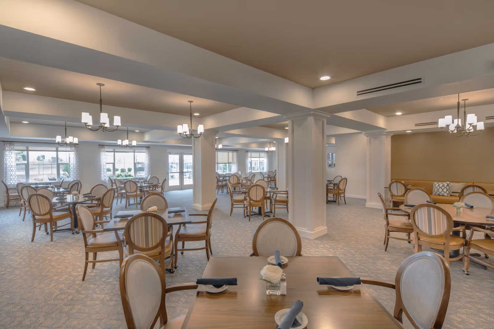 A spacious dining room with multiple round wooden tables and cushioned chairs arranged neatly. The room features large windows allowing natural light, chandeliers hanging from the ceiling, and a carpeted floor. The walls are painted in neutral tones with white columns dividing the space.