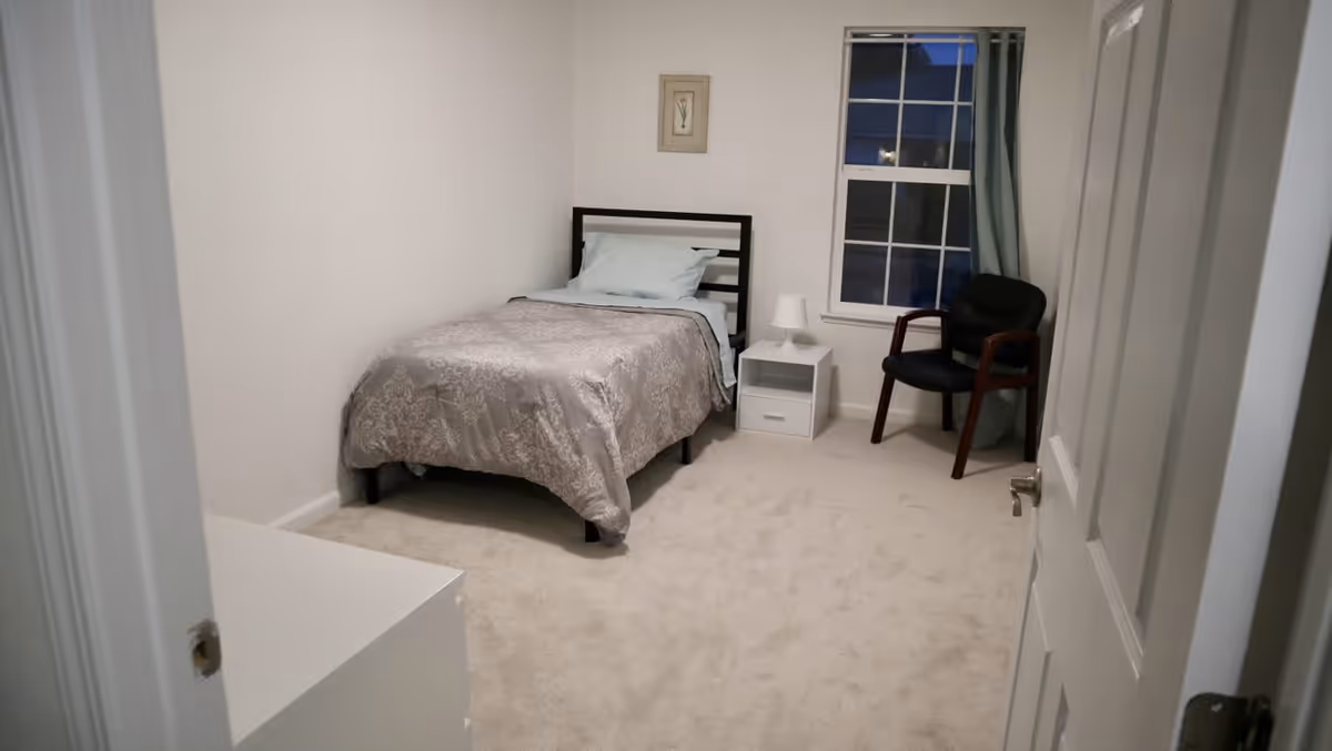 A small, simple bedroom with a single bed covered in a gray patterned comforter and light blue pillow. Next to the bed is a white nightstand with a small white lamp. A black chair with wooden armrests is placed near a window with blue curtains. The room has beige carpet and white walls.