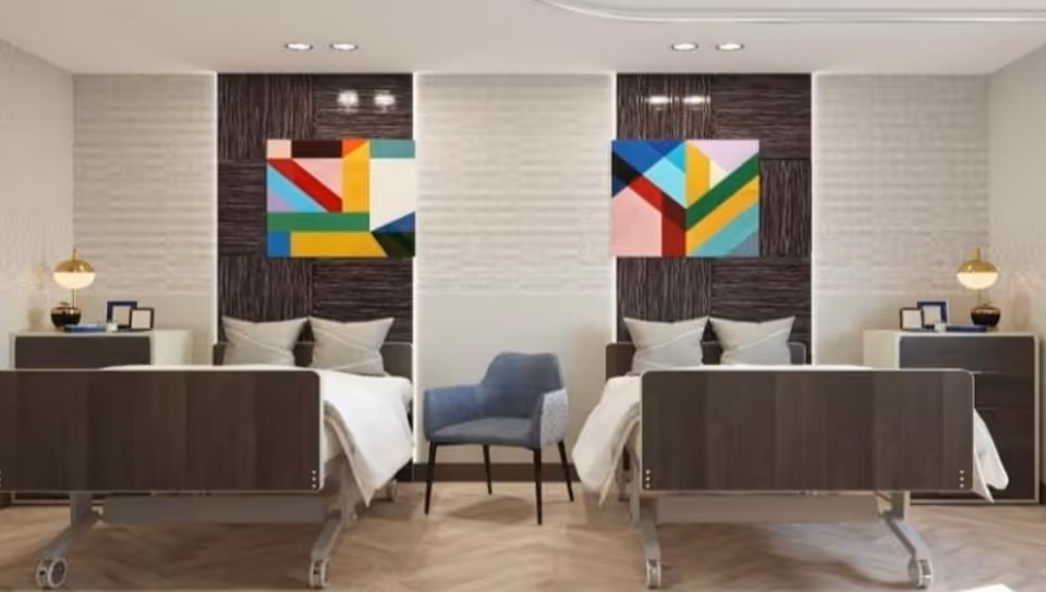 A modern, clean room with two hospital-style beds separated by a partial wall. Each bed has white bedding and pillows, a bedside table with a lamp, and colorful abstract artwork above the headboard. A blue upholstered chair is placed between the beds.