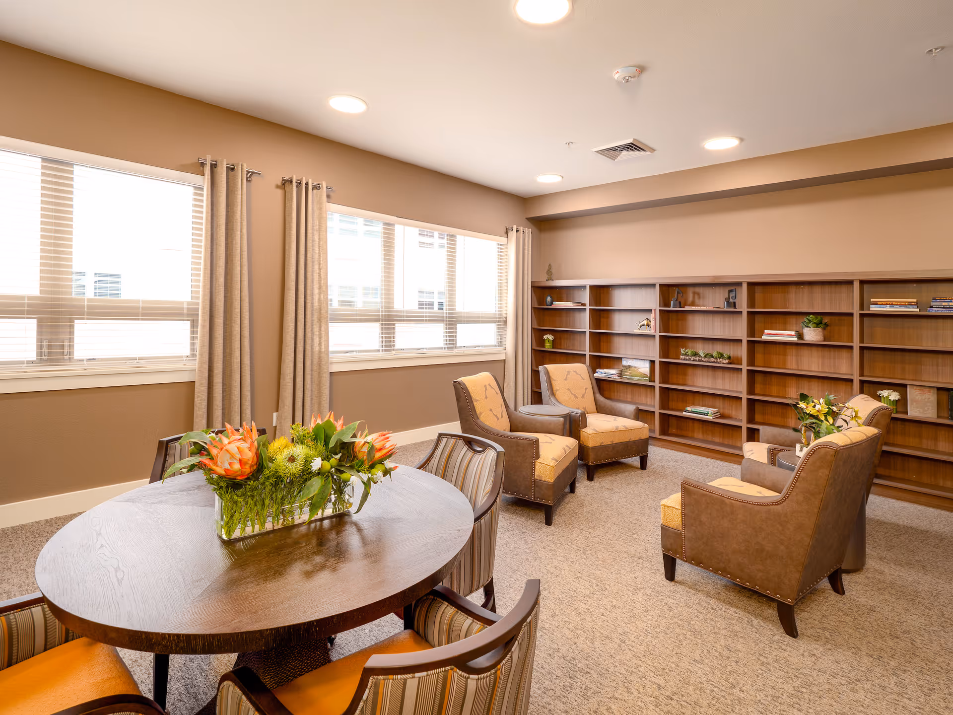 A cozy senior living community room with a round wooden table adorned with a floral centerpiece, surrounded by four striped chairs. The room features four upholstered armchairs arranged near a large wooden bookshelf with some books and decorative plants. Large windows with beige curtains allow natural light to fill the space, creating a warm and inviting atmosphere.