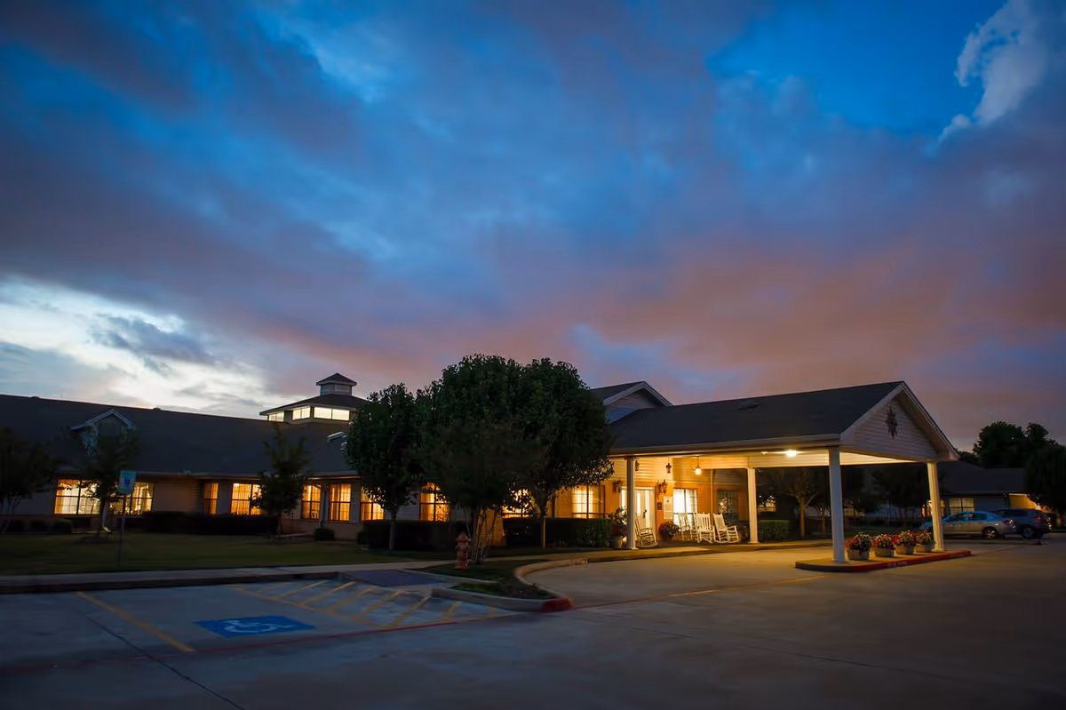 Exterior of a single-story senior living facility at dusk with a lit covered entrance and rocking chairs.
