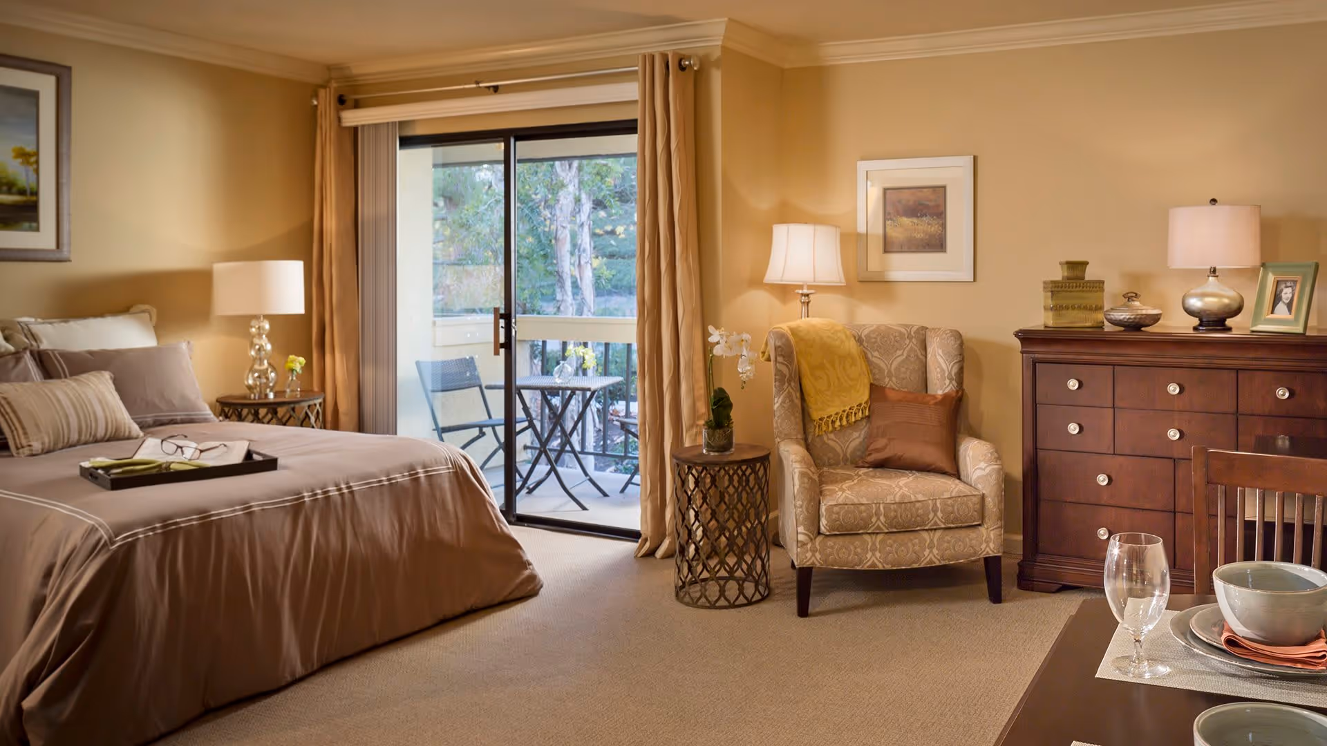 A cozy bedroom with a large bed dressed in brown bedding, a bedside table with a lamp, an armchair with a yellow throw and brown pillow, a wooden dresser with decorative items and a framed photo, and a sliding glass door leading to a small outdoor patio with a table and two chairs.