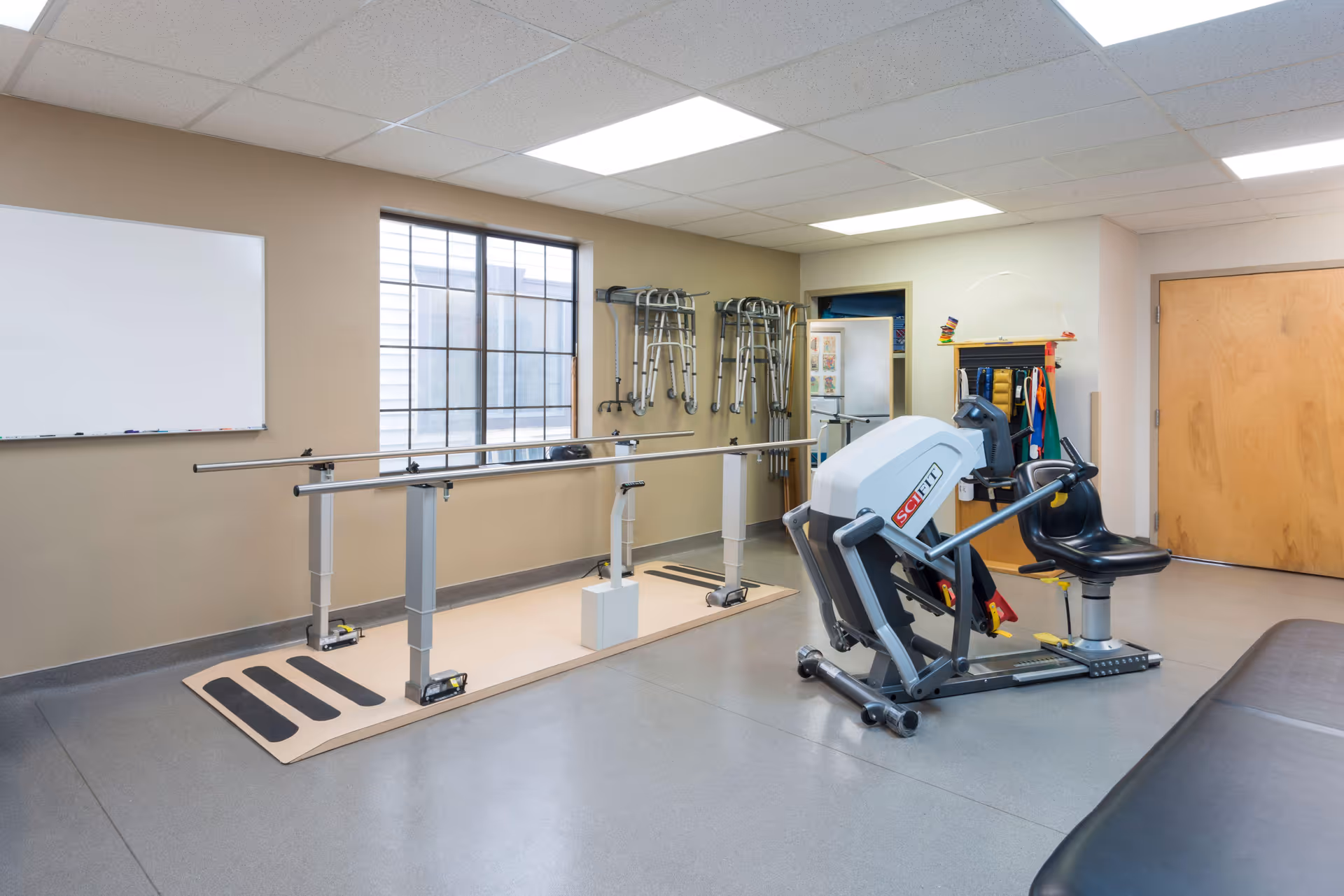 A rehabilitation or physical therapy room with parallel bars for walking exercises, a seated exercise machine, several walkers hanging on the wall, a whiteboard, and a wooden door. The room has a window letting in natural light and a clean, organized appearance.