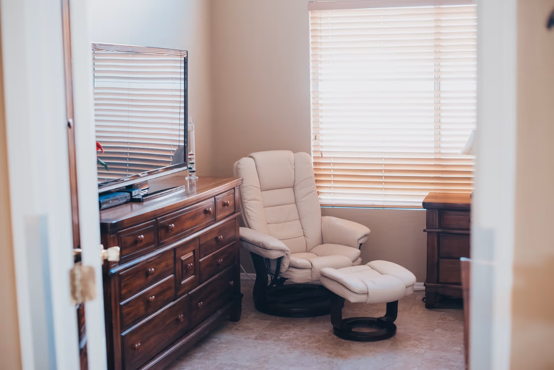 A cozy room with a cream-colored recliner chair and matching footrest positioned near a window with closed blinds. There is a wooden dresser with a flat-screen TV on top and a small wooden nightstand beside the chair. The room has light-colored walls and flooring.