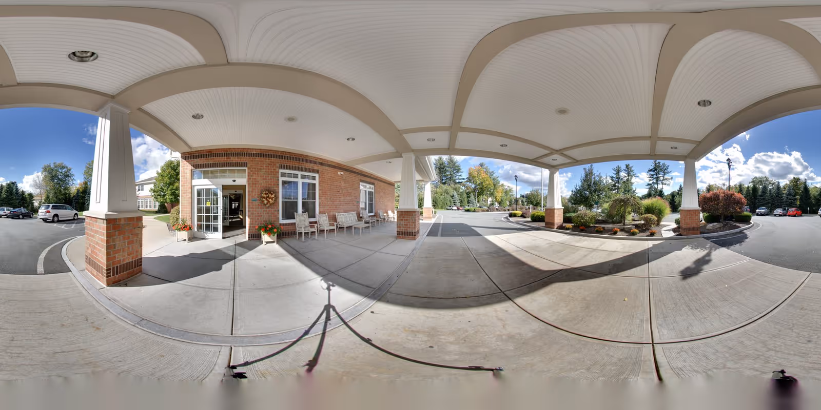 Covered entrance area of Peregrine Senior Living at Delmar Place with brick pillars and white ceiling beams. There are chairs and potted plants near the entrance door. The driveway and parking area surround the entrance, with trees and landscaping visible under a partly cloudy blue sky.
