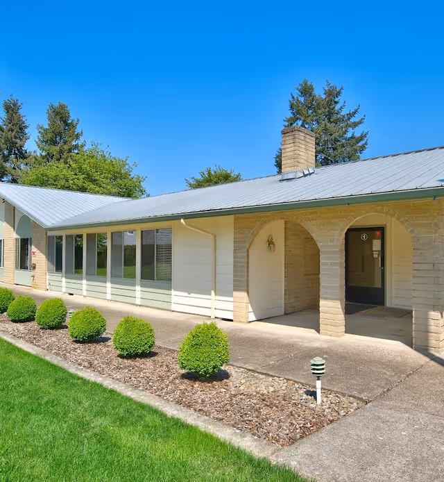 Exterior view of a single-story building with a metal roof and arched entryways. The building is surrounded by neatly trimmed bushes, a mulched garden bed, and a well-maintained lawn under a clear blue sky.