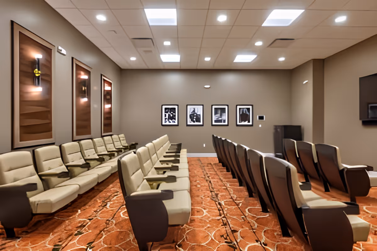 Carpeted community media room with rows of cushioned theater-style seats facing a blank front wall and framed artwork.