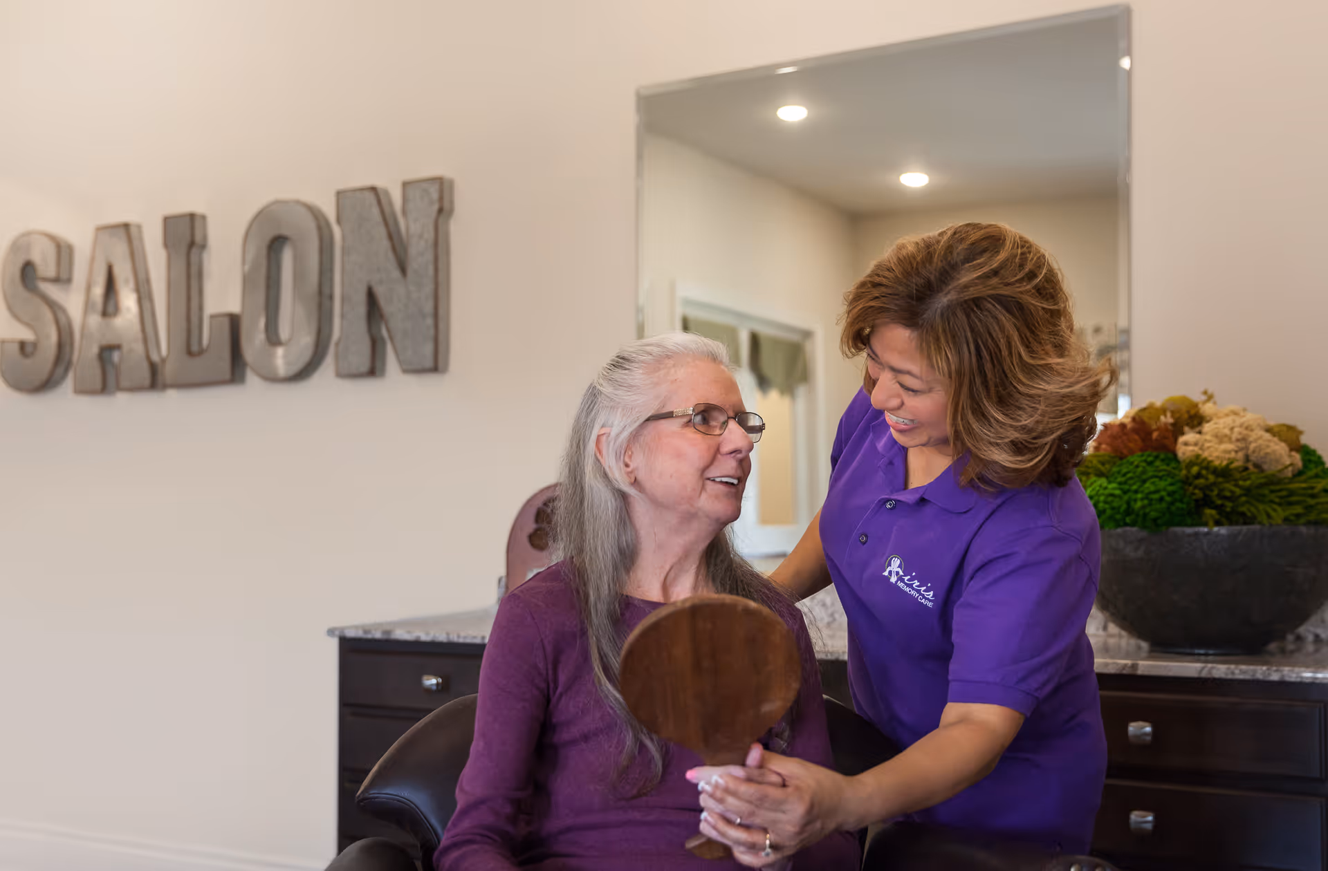 A senior woman with long gray hair and glasses sits in a salon chair holding a hand mirror, smiling at a caregiver in a purple polo shirt who is standing beside her and smiling back. Behind them is a large mirror and a decorative bowl with green and beige floral arrangements. The word 'SALON' is displayed on the wall in large metallic letters.