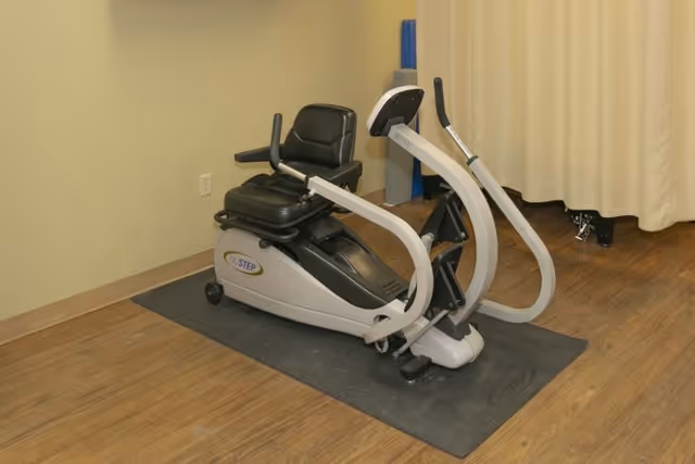 A recumbent cross-trainer exercise machine on a rubber mat in a small interior room with wood flooring and a curtained wall.