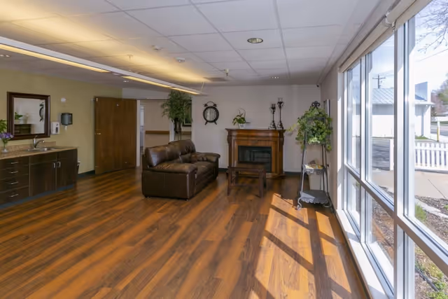 Sunlit common living area with a leather sofa, fireplace, hardwood floors, and large windows.
