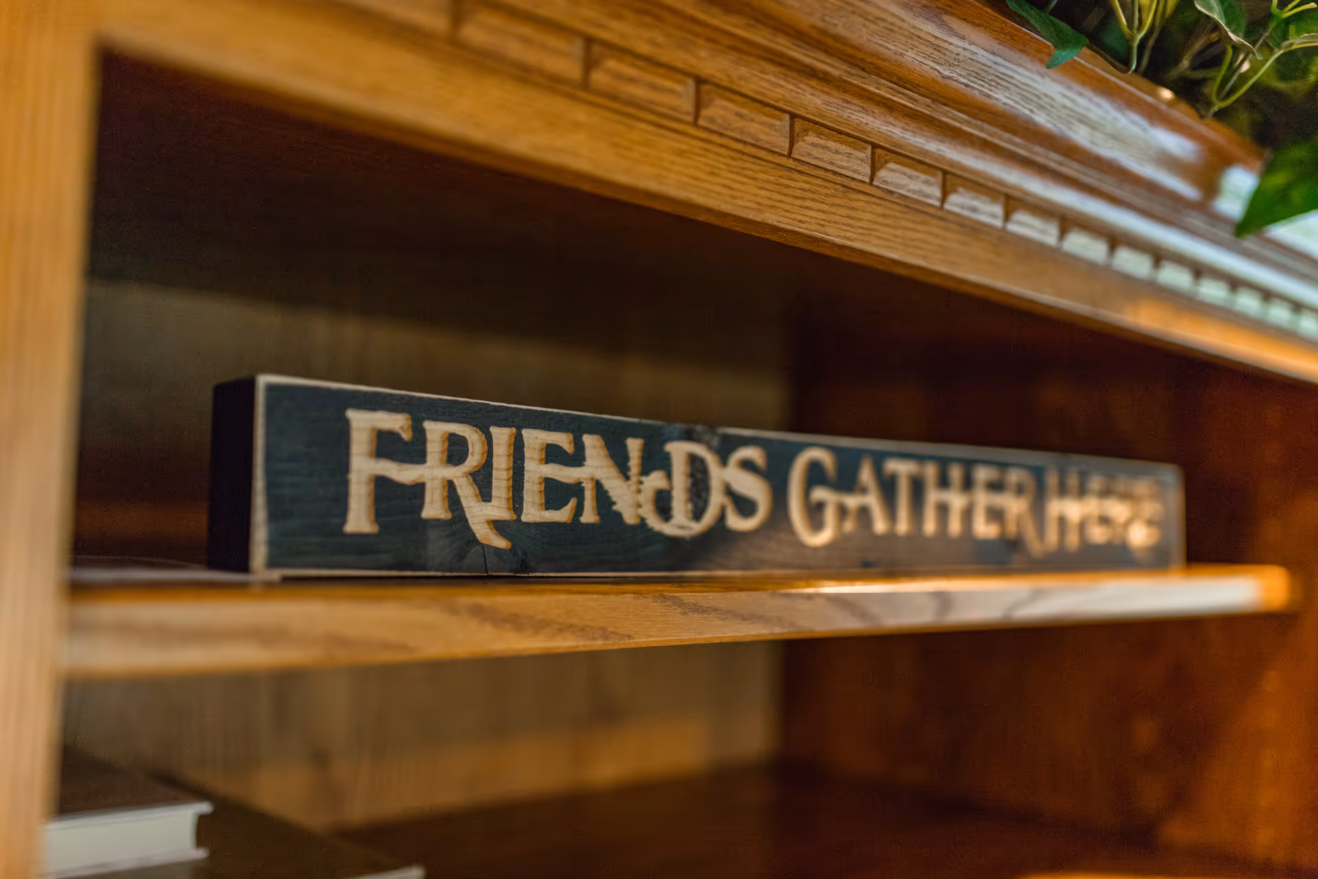 A wooden decorative sign on a wooden shelf that reads 'FRIENDS GATHER HERE' in carved letters.