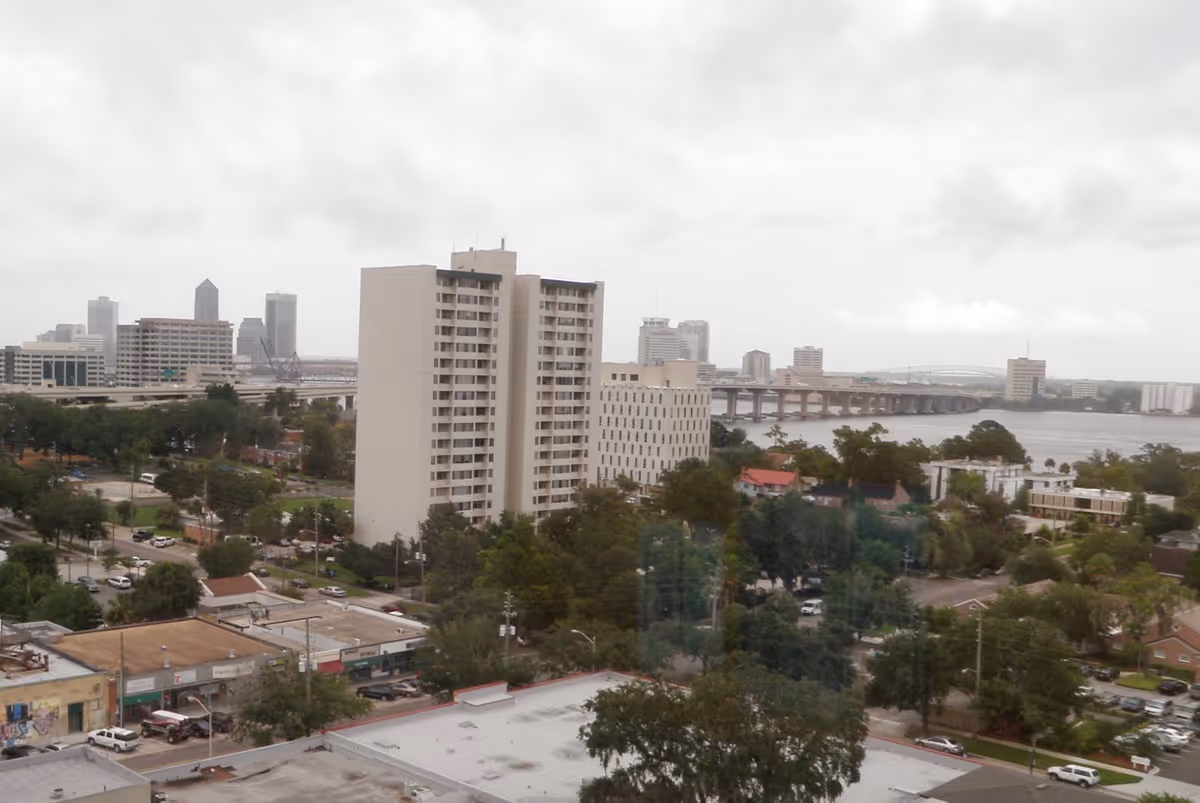 View of Riverside Presbyterian Apartments, a tall residential building near a river with a bridge in the background and a city skyline visible in the distance under a cloudy sky.