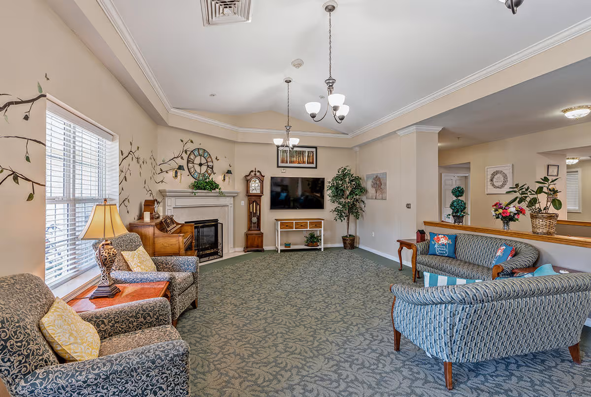A cozy senior living facility common area with patterned armchairs and sofas arranged around a carpeted floor. The room features a fireplace with a large clock above it, a grandfather clock, a wall-mounted TV, and decorative plants. The walls are adorned with nature-themed artwork and wall decals of tree branches. Ceiling lights hang from a tray ceiling, and a window with blinds allows natural light to enter.