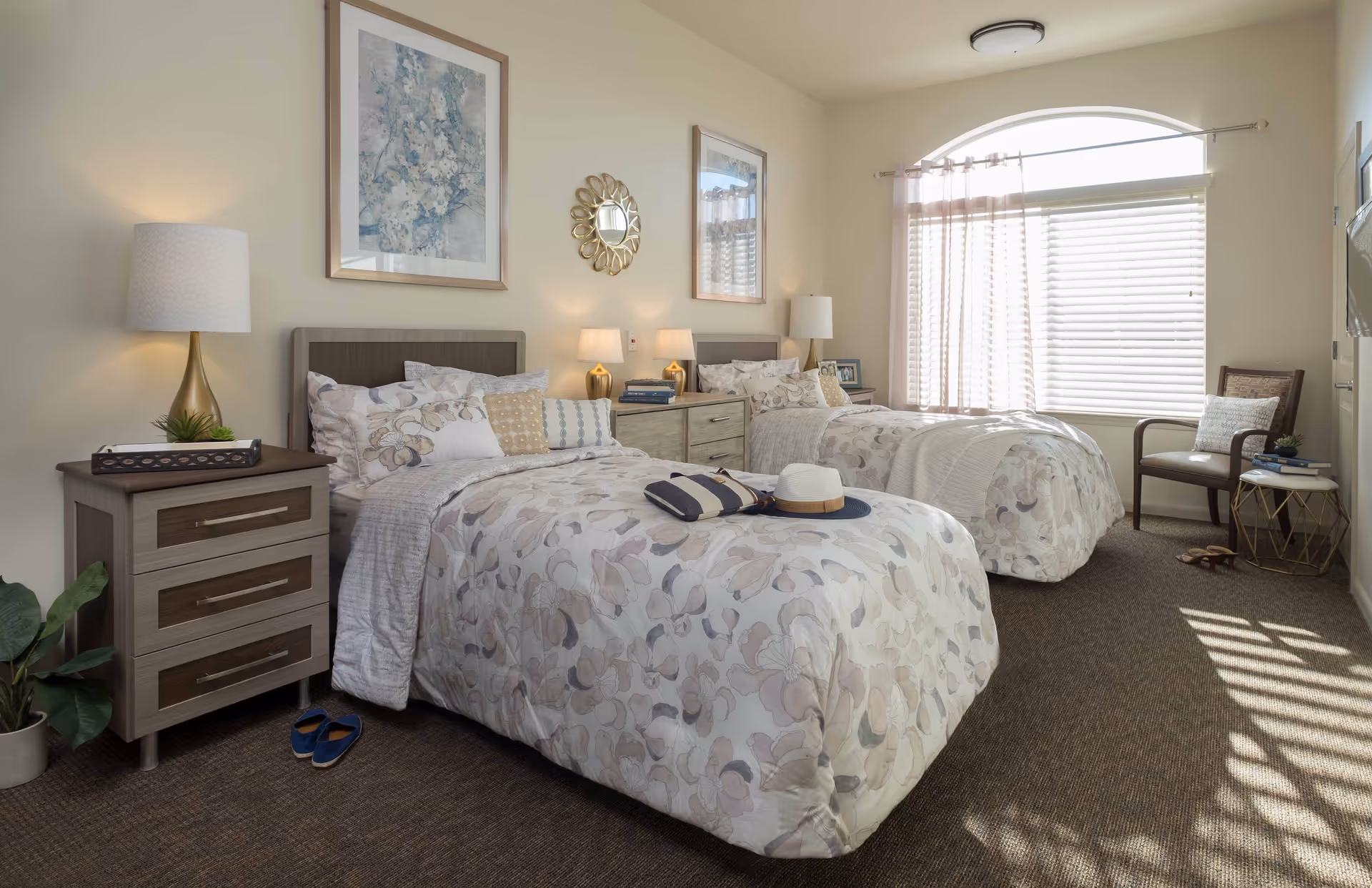 Well-lit twin bedroom with two patterned beds, nightstands, lamps, and a large arched window.