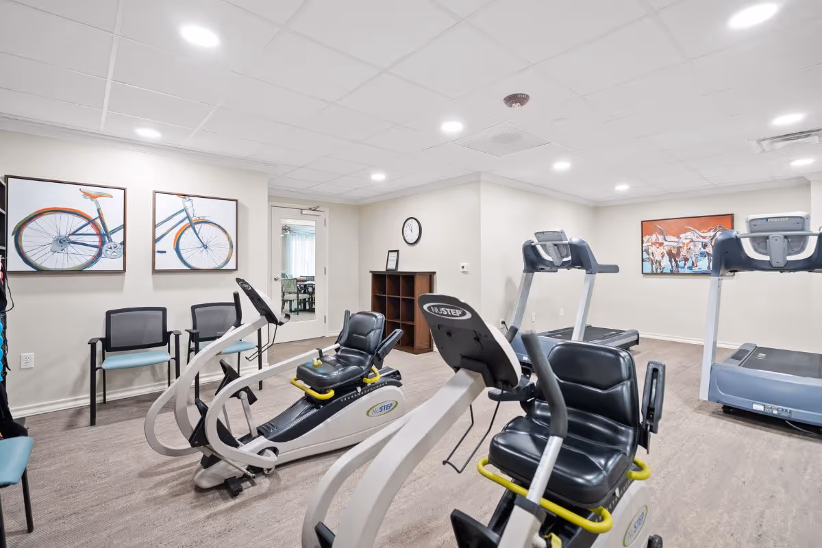 Bright fitness room with recumbent exercise machines, treadmills, chairs, and framed artwork on the walls.