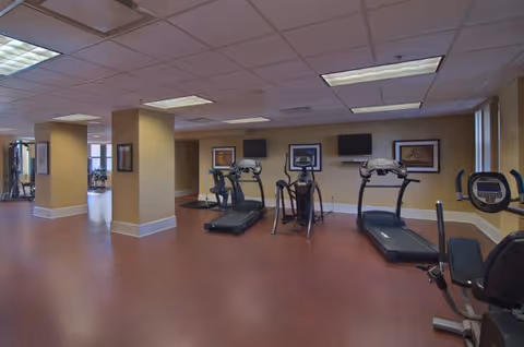 Interior view of a fitness room with exercise equipment including treadmills, stationary bikes, and elliptical machines. The room has beige walls, framed pictures, and a ceiling with fluorescent lights.