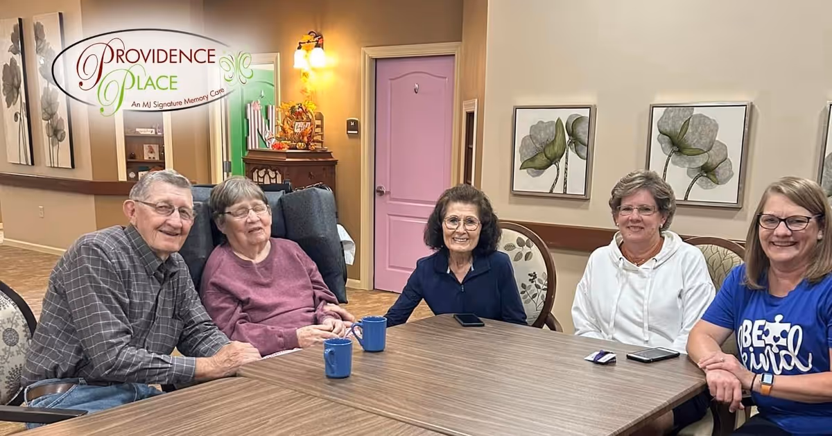 Five elderly individuals sitting around a wooden table in a cozy room with beige walls, floral paintings, and a pink door in the background. Two blue mugs are on the table, and the group is smiling and appears to be enjoying each other's company.