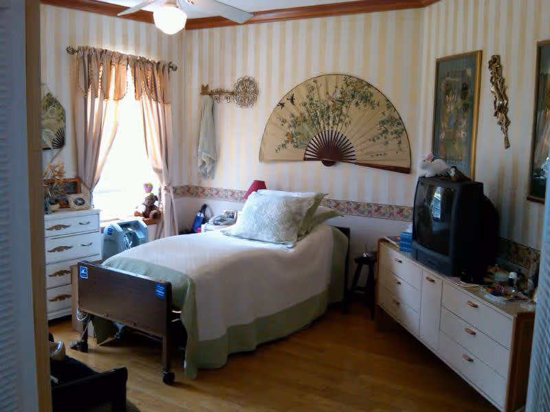 A cozy bedroom with a single bed covered in white and green bedding. The room has striped wallpaper with a floral border and a large decorative fan hanging above the bed. There is a window with curtains on the left side, a dresser with a mirror and various items, and a TV on a long dresser on the right side. The floor is wooden, and the room is softly lit by natural light from the window.