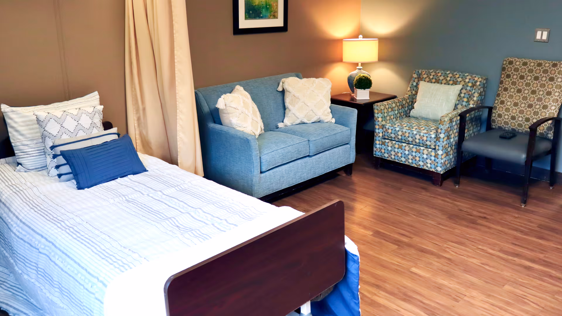 A cozy room featuring a single bed with multiple pillows, a blue loveseat with two white cushions, two patterned armchairs, a wooden side table with a lamp and a small plant, and wooden flooring.