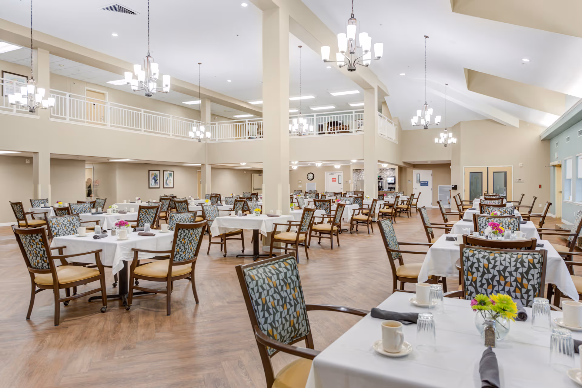 Spacious, well-lit dining room with multiple tables set with white tablecloths and patterned chairs in a senior living facility.