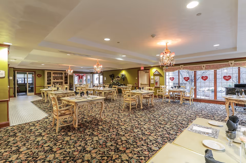 Spacious dining room with multiple set tables and chairs, chandeliers, patterned carpet, and large windows decorated with heart ornaments.
