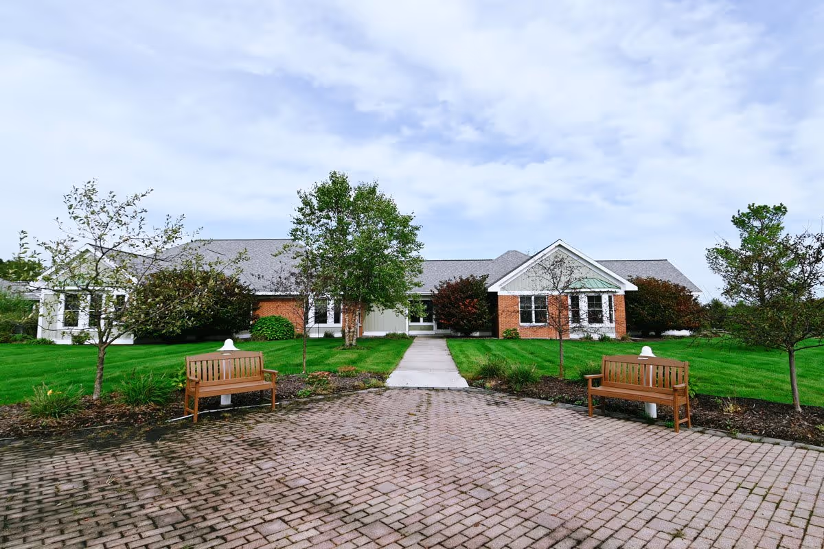 Front exterior view of The Cortland Holland Meadows building with a paved walkway leading to the entrance, flanked by two wooden benches and small trees on a well-maintained lawn under a partly cloudy sky.