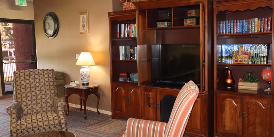 A cozy living room area in an assisted living facility featuring a patterned armchair and a striped armchair facing a wooden entertainment center with a flat-screen TV. The entertainment center also has shelves filled with books and decorative items. A small wooden side table with a lamp and a clock on the wall are visible, along with a glass door leading outside.