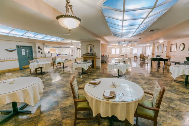 Bright communal dining room with round tables covered in white linens, chairs, and skylight ceiling panels.