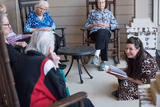 A group of elderly women sitting on rocking chairs on a covered porch or patio, engaged in conversation with a younger woman who is kneeling on the floor holding an open book and smiling.