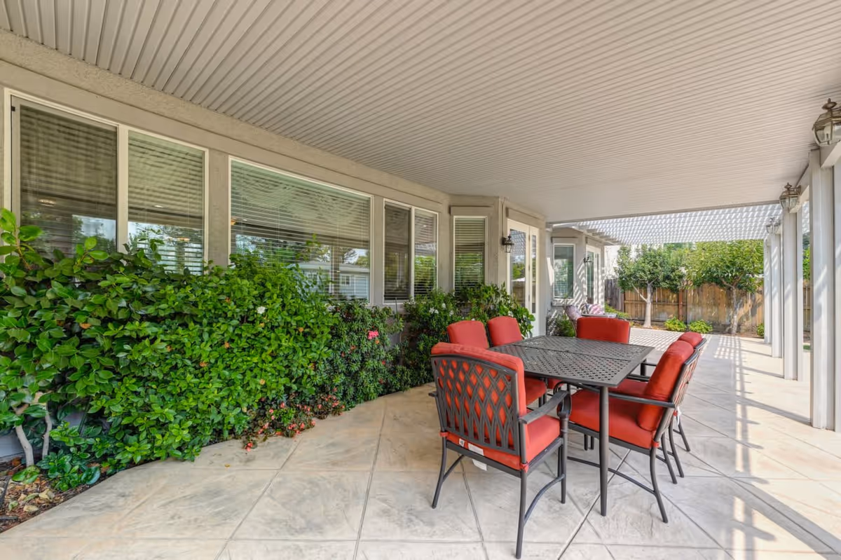 Covered outdoor patio area with a black metal table and six chairs with red cushions. The patio has a white ceiling and is adjacent to a building with large windows and green bushes along the wall. In the background, there is a wooden fence and some trees.