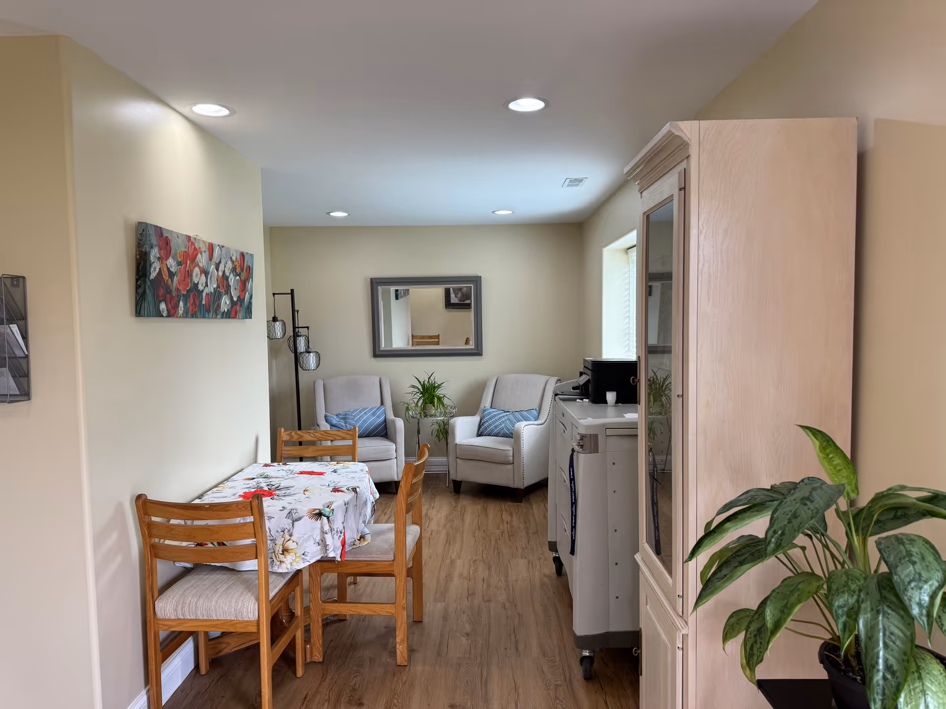 A cozy interior room with light beige walls and wood flooring. There is a small wooden dining table with a floral tablecloth and four chairs on the left side. At the back of the room, two cushioned armchairs with blue patterned pillows are placed near a window, with a small plant on a stand between them. A large mirror hangs on the back wall above the chairs. On the right side, there is a wooden cabinet and a white rolling cart with a coffee maker on top. A green potted plant is visible in the foreground on the right.