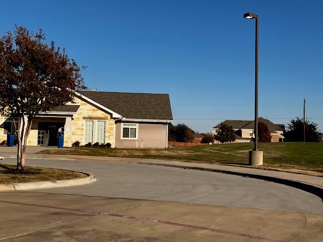 Exterior view of a single-story building with a stone and beige facade, a tree with autumn leaves on the left, a curved driveway in front, and a tall street lamp on the right under a clear blue sky.