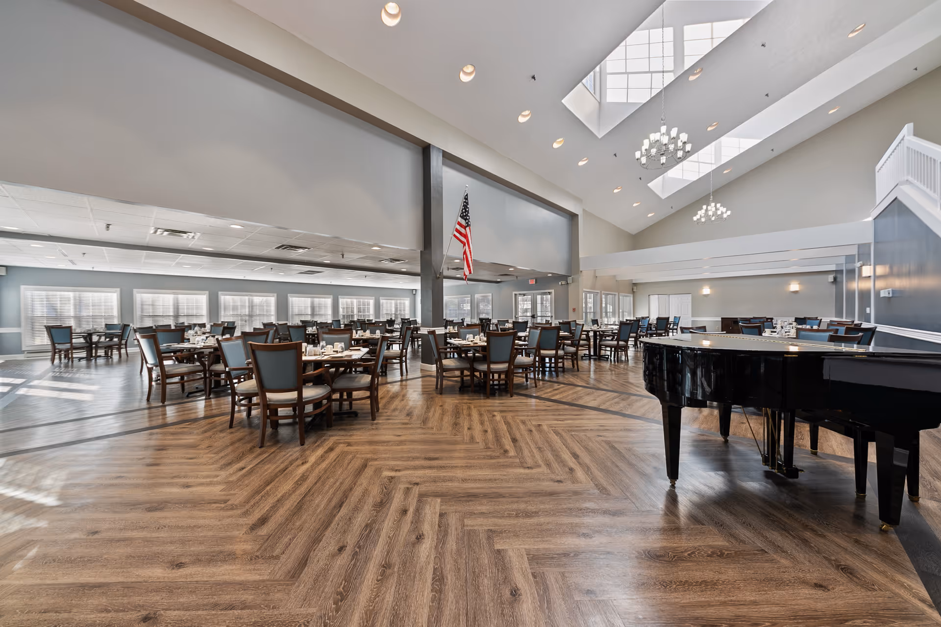 Spacious dining room with multiple tables and chairs arranged neatly. The room features large windows allowing natural light, a high ceiling with skylights and chandeliers, and a black grand piano on the right side. An American flag is displayed near the center pillar.