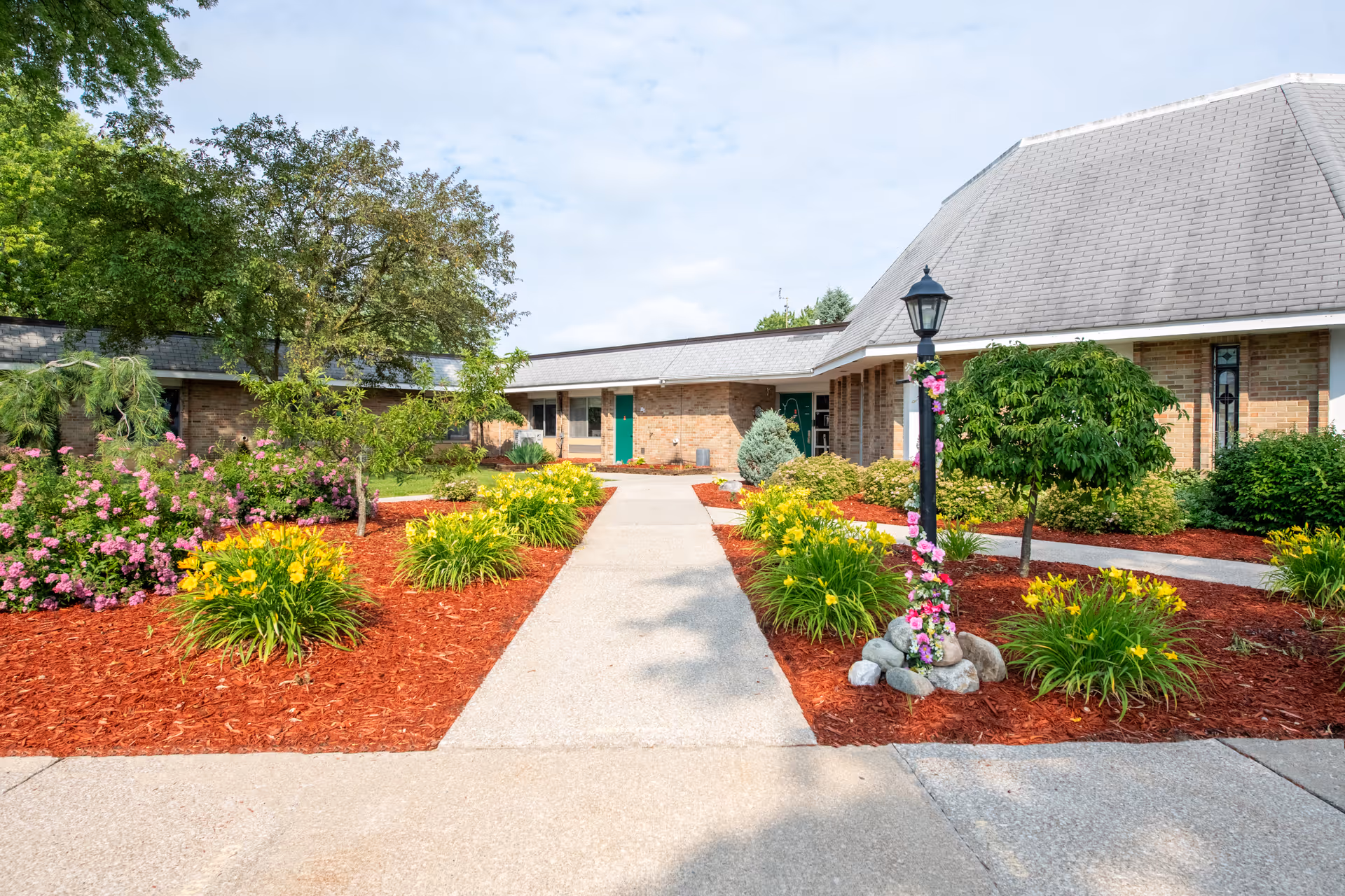 Concrete walkway leading to the front entrance of a single-story brick senior living building with landscaped flower beds and a lamppost.
