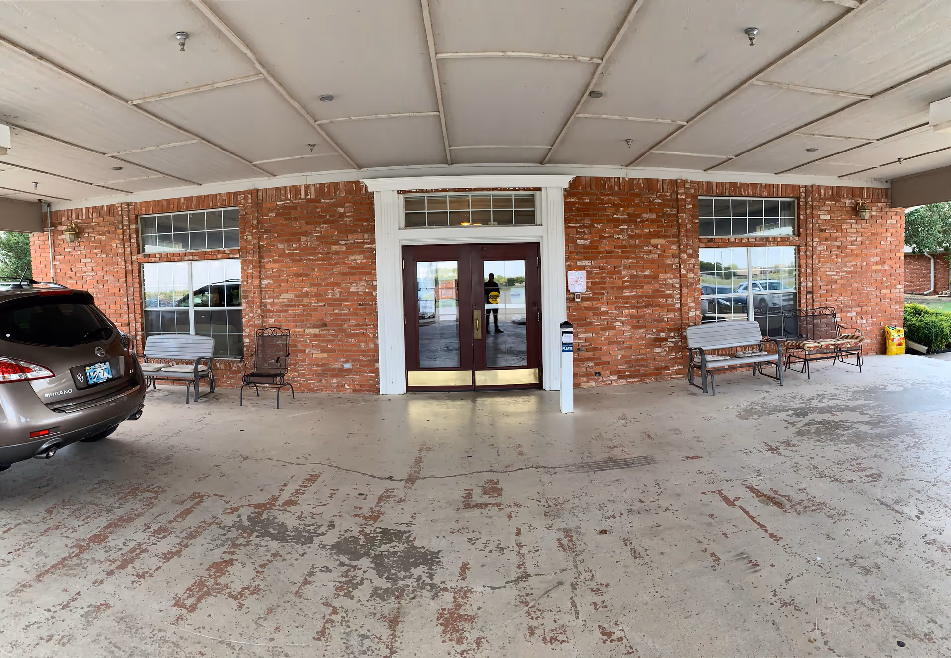 Covered brick entrance with double glass doors, benches, and a parked car under a porte-cochere.