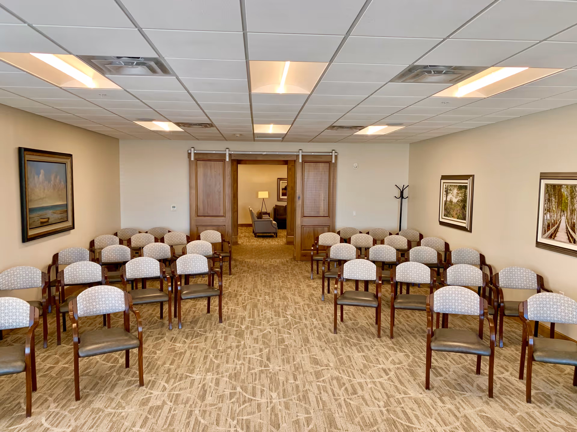 A conference or meeting room with multiple rows of wooden chairs with cushioned seats and backs arranged facing forward. The room has beige walls adorned with framed landscape paintings and a carpeted floor. At the far end, there are double wooden sliding doors leading to another room with armchairs and a lamp.