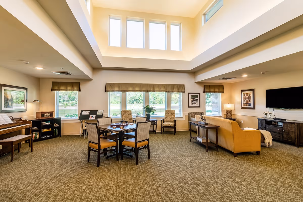 A spacious, well-lit common living area with large windows and high ceilings. The room features a yellow sofa facing a wall-mounted flat-screen TV, a wooden console table, a round table with four chairs, and additional seating near the windows. There is a piano and bookshelf on the left side of the room, and framed artwork on the walls. The carpet is a neutral tone, and the room has a warm, inviting atmosphere.