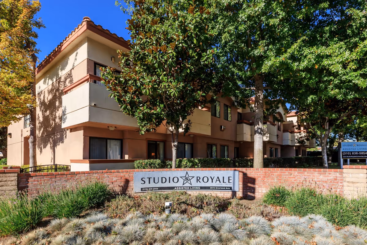 Exterior view of Studio Royale assisted living facility showing a two-story building with balconies, surrounded by trees and landscaping. A brick wall in front displays a sign with the facility's name and contact information.