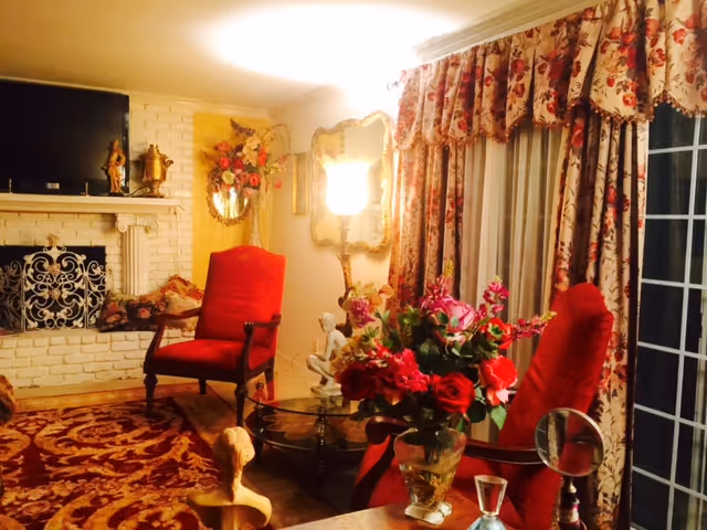 A cozy living room with two red upholstered armchairs, a glass coffee table with a vase of red and pink flowers, floral curtains covering a large window or sliding door, a white brick fireplace with decorative items on the mantel, a wall mirror, and warm lighting.
