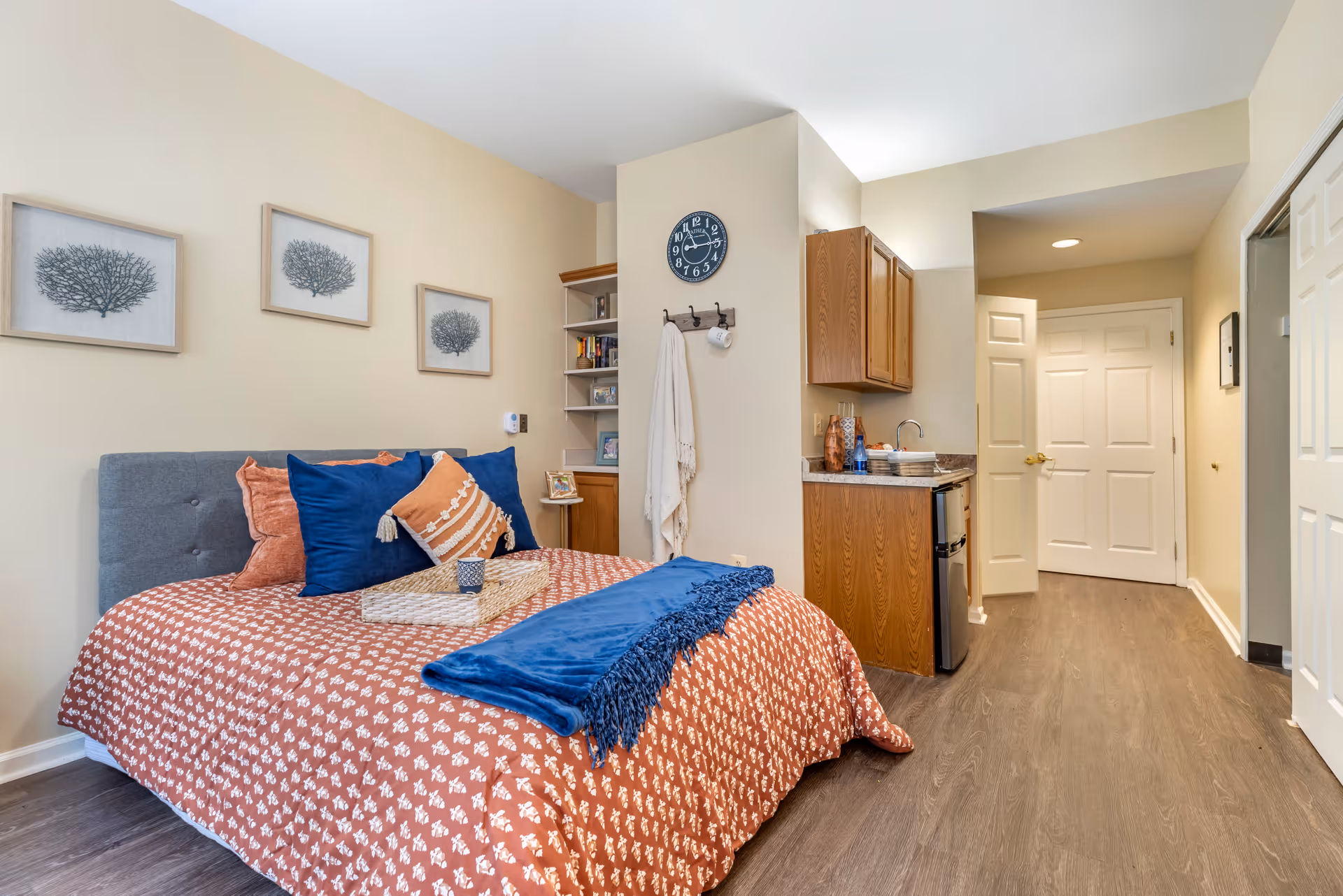 A cozy bedroom in a senior living facility with a bed covered in a rust-colored patterned bedspread and blue and orange pillows. Above the bed are three framed coral prints. To the right of the bed is a small kitchenette with wooden cabinets, a mini fridge, and a countertop with a sink. A clock and a wall hook with a hanging towel and mug are mounted on the wall between the bed and kitchenette. The room has wood flooring and neutral-colored walls.