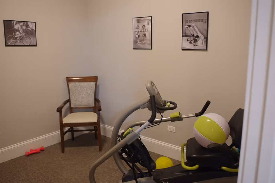 Small interior exercise room with a stationary exercise machine, a chair, a pink dumbbell, and framed photos on the wall.