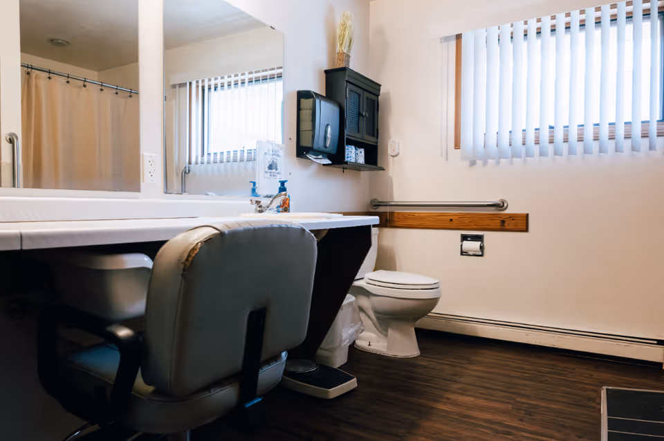 A bathroom with a white toilet, a large mirror above a countertop with a sink, a padded chair with worn edges, a wall-mounted cabinet, a paper towel dispenser, and a window with vertical blinds. The floor is dark wood, and there is a grab bar on the wall next to the toilet.