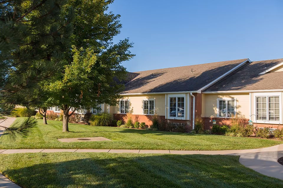 Exterior view of a single-story residential building with beige siding and brick accents, surrounded by green grass, trees, and a paved walkway under a clear blue sky.