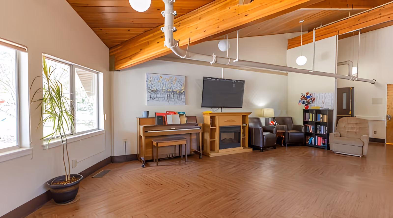 A cozy common area with a wooden piano, a flat-screen TV mounted above a wooden electric fireplace, two leather armchairs, a beige upholstered armchair, a bookshelf filled with books, a potted plant near large windows, and a wooden ceiling with exposed beams and hanging round lights.