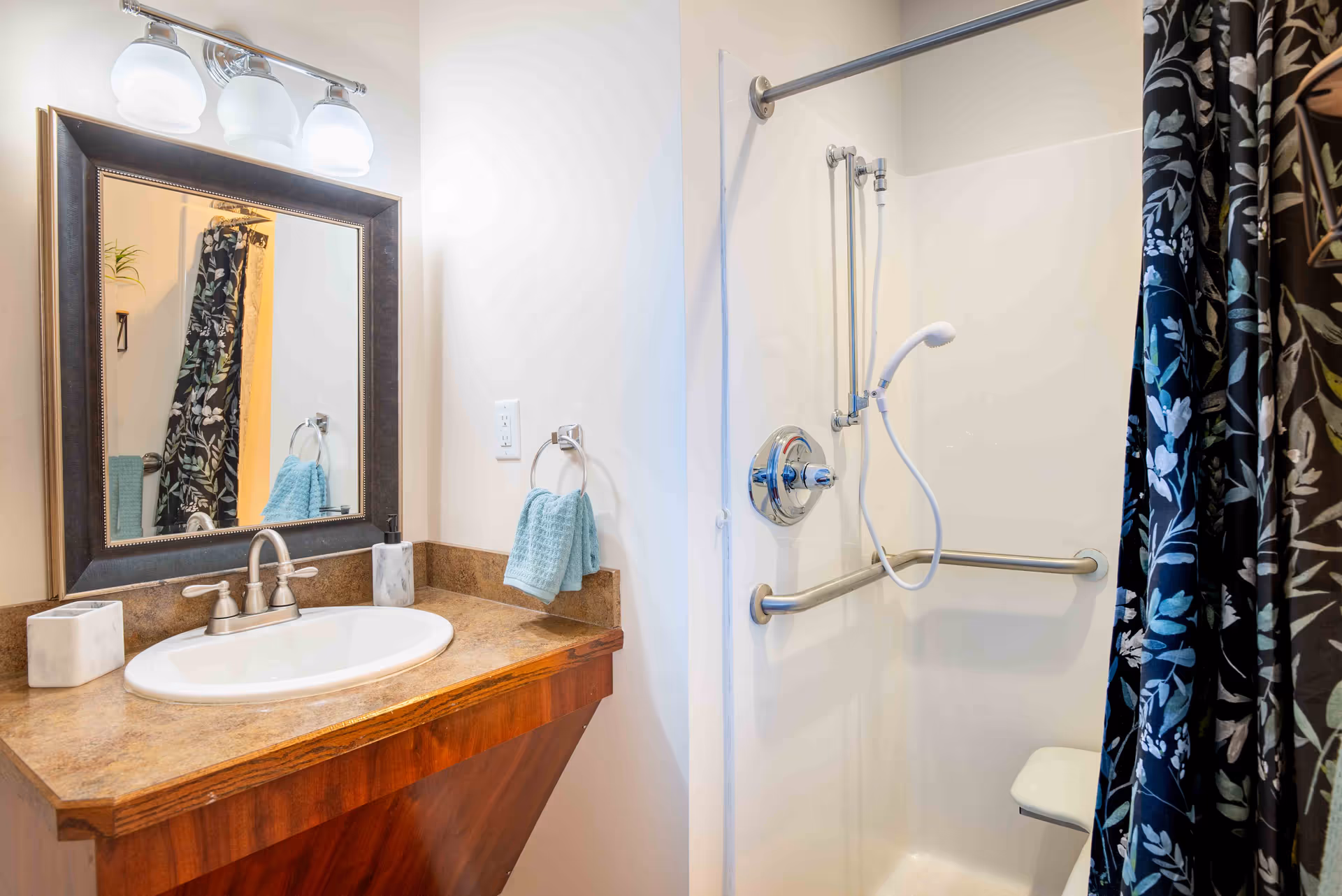 A bathroom featuring a wooden vanity with a brown countertop, a white sink with silver faucet, a rectangular mirror with a dark frame, and three light fixtures above the mirror. To the right is a shower area with a white wall, a handheld showerhead, a horizontal grab bar, and a floral-patterned shower curtain partially drawn.