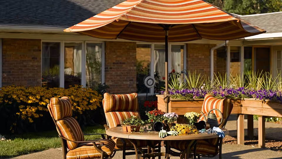 Outdoor patio area with a round table surrounded by four cushioned chairs with orange and beige striped upholstery. A large matching striped umbrella provides shade. The table has small potted plants, gardening gloves, and tools on it. Behind the patio, there are raised garden beds with purple flowers and a brick building with windows.